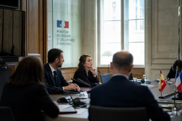 Table de négociation internationale à Paris avec micros, dossiers fermés et silhouettes anonymes sous une lumière claire.