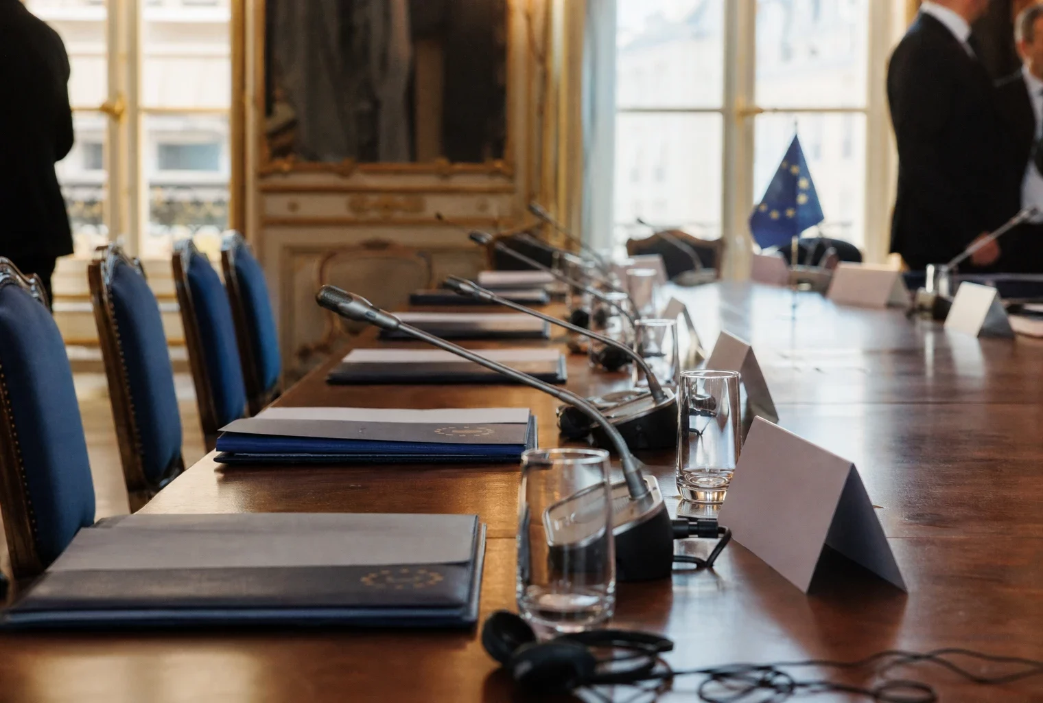 Table de négociation européenne avec dossiers, casques et drapeaux miniatures dans une salle institutionnelle lumineuse.