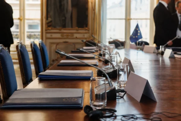 Table de négociation européenne avec dossiers, casques et drapeaux miniatures dans une salle institutionnelle lumineuse.