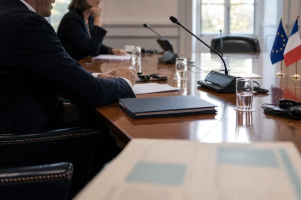 Table de négociation institutionnelle avec micros, dossiers fermés et silhouettes anonymes sous une lumière claire.
