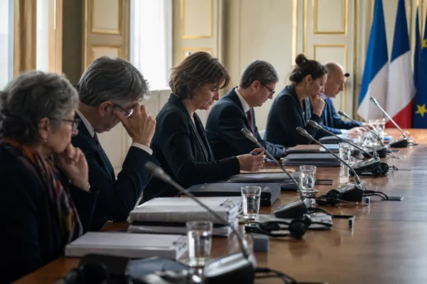 Table de négociation institutionnelle avec dossiers fermés, micros et silhouettes anonymes dans une lumière naturelle claire.