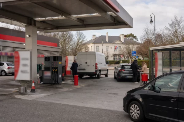 Station-service en périphérie française avec voitures civiles et clients anonymes, dans une ambiance de reportage claire.