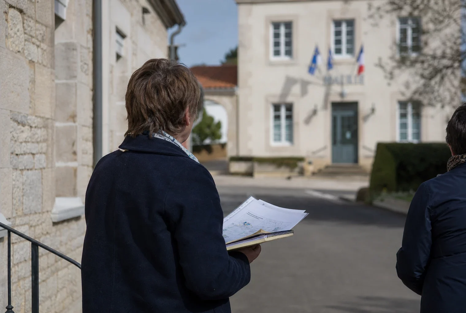 Un élu local de dos devant la mairie d’un village vendéen, dossier en main, dans une ambiance de campagne discrète.