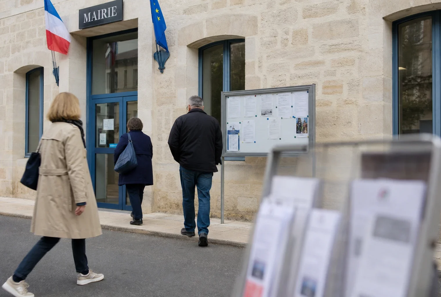 Des anonymes se rendent devant une mairie de ville moyenne, sous une lumière naturelle claire et apaisée.