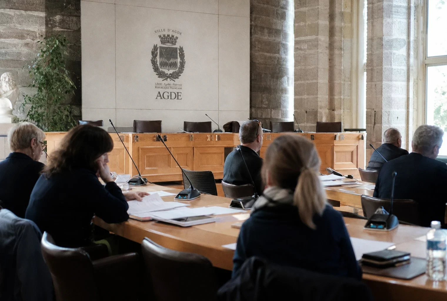 Salle municipale d’Agde en plan large avec micros, dossiers flous et chaise vide, ambiance de reportage