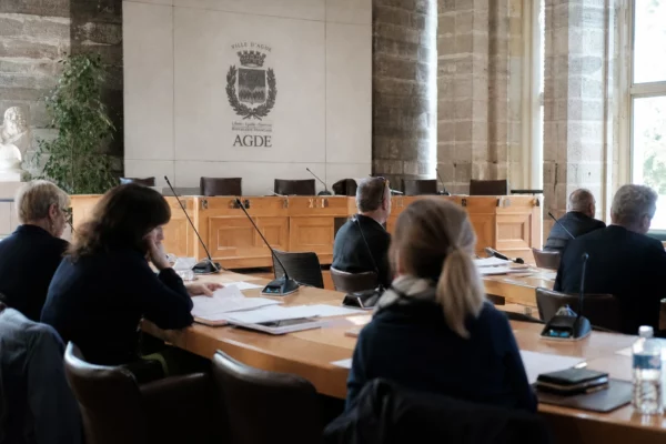 Salle municipale d’Agde en plan large avec micros, dossiers flous et chaise vide, ambiance de reportage