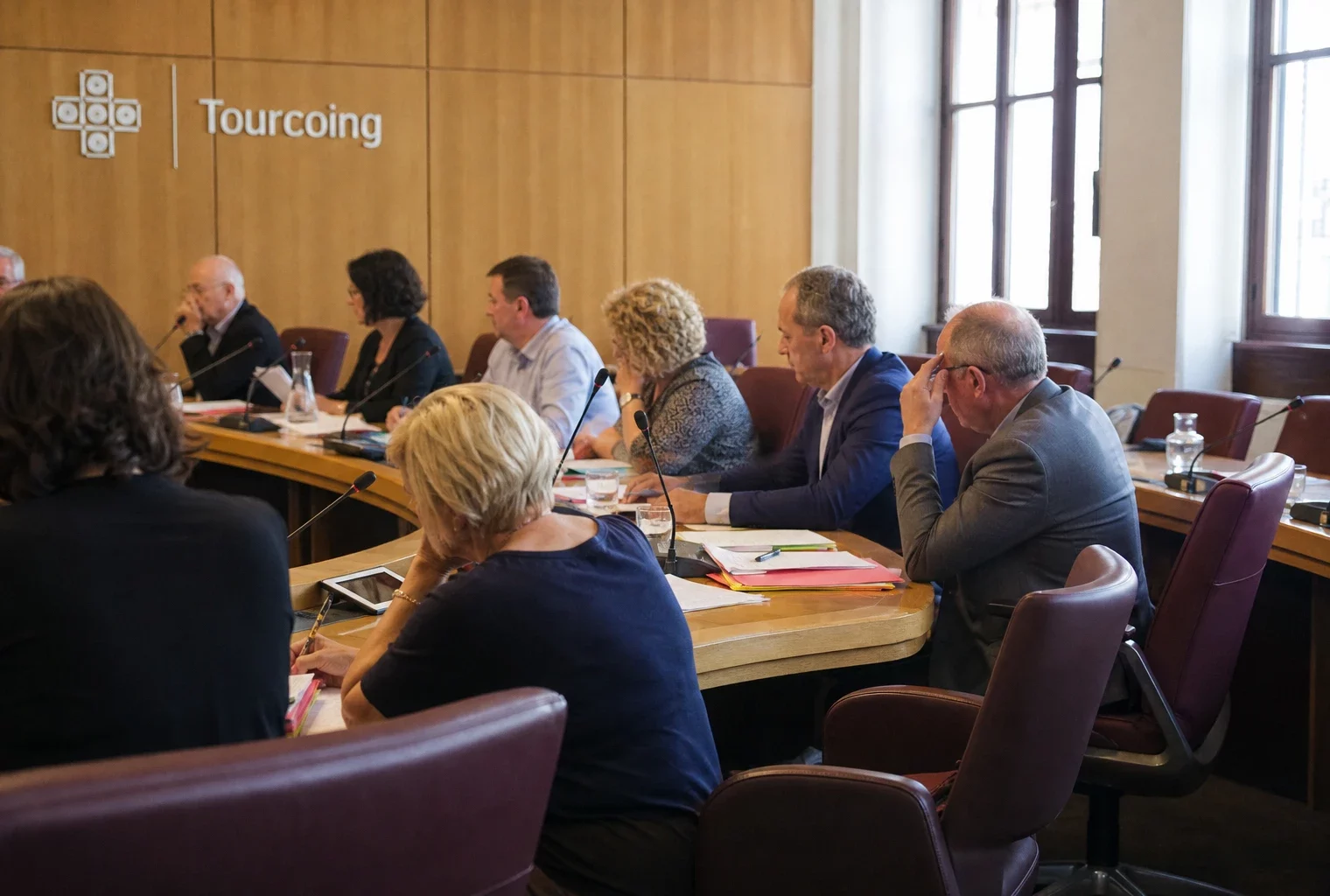 Salle municipale française avec élus anonymes, micros, dossiers flous et une chaise vide autour d’une table
