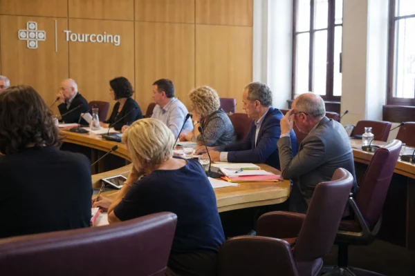 Salle municipale française avec élus anonymes, micros, dossiers flous et une chaise vide autour d’une table