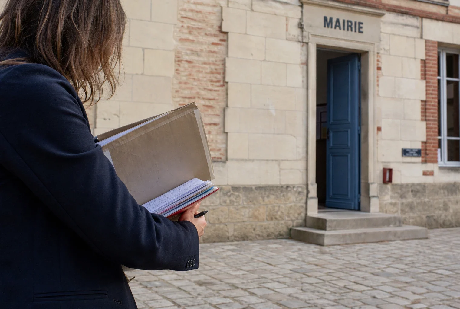 Un élu local anonyme tient un dossier devant une mairie française, dans une scène documentaire lumineuse.