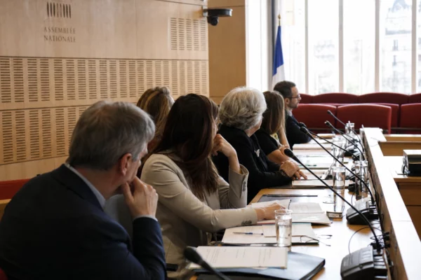Audition institutionnelle dans une salle parlementaire française, avec micros, dossiers flous et silhouettes anonymes