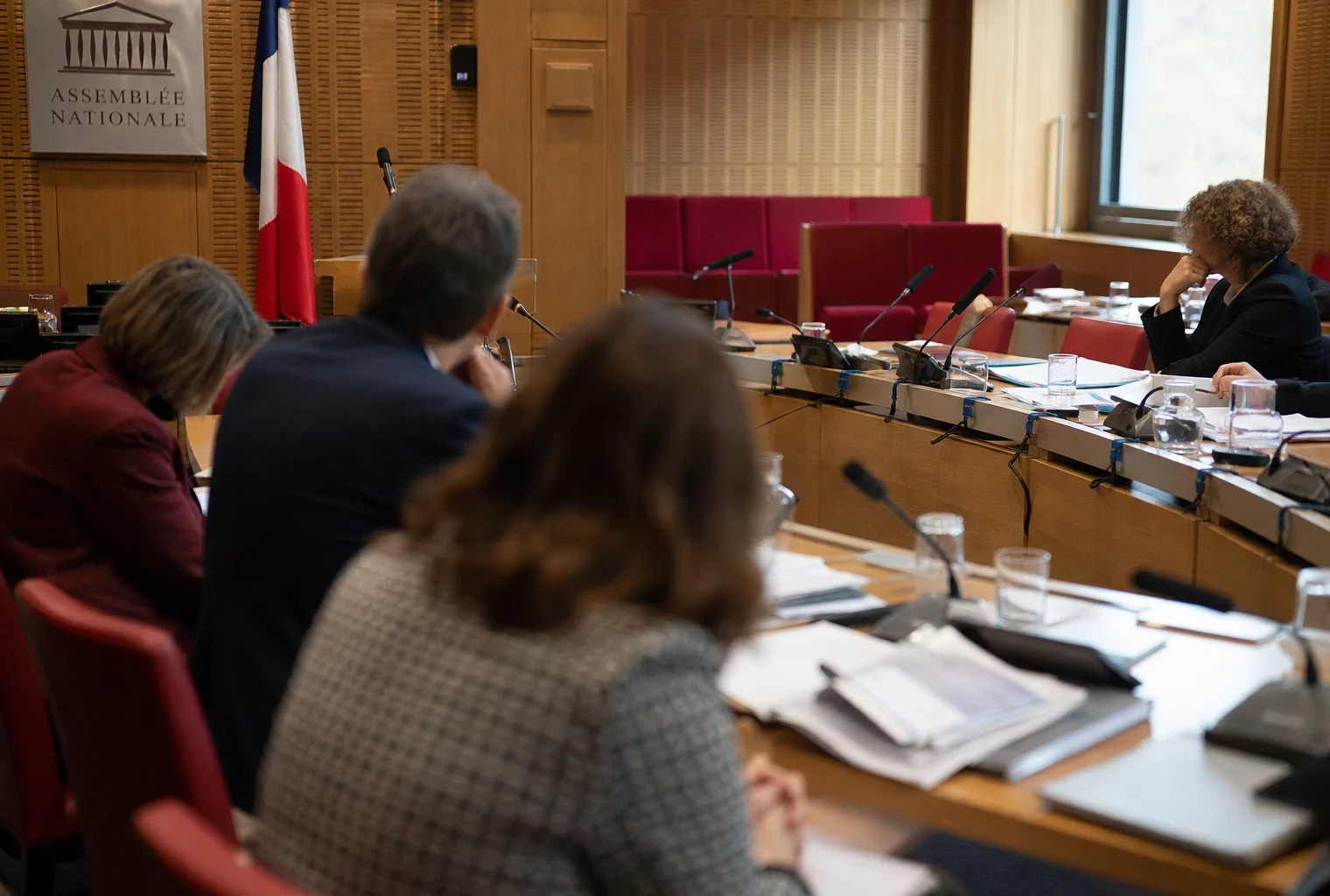 Audition parlementaire en salle de commission avec micros, dossiers ouverts et silhouettes anonymes sous une lumière claire.