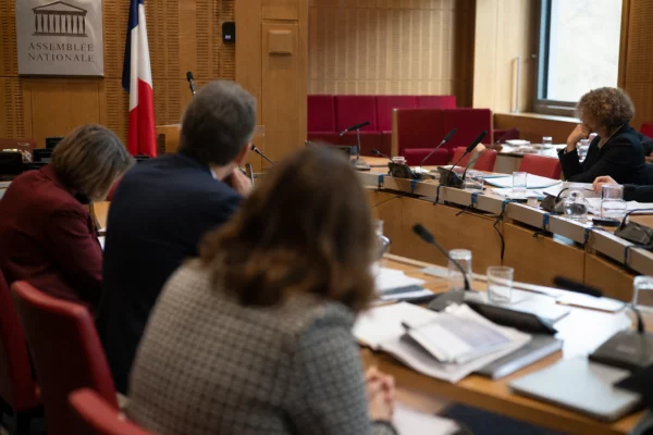 Audition parlementaire en salle de commission avec micros, dossiers ouverts et silhouettes anonymes sous une lumière claire.