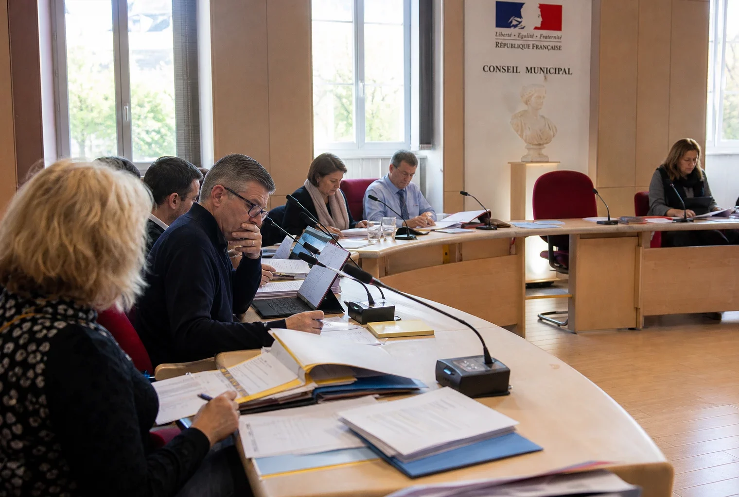 Salle de conseil municipal française avec une chaise vide, des micros et des dossiers budgétaires sur une table