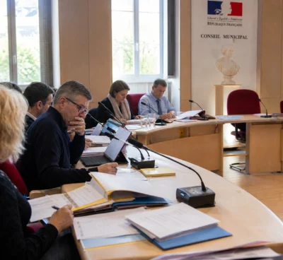 Salle de conseil municipal française avec une chaise vide, des micros et des dossiers budgétaires sur une table