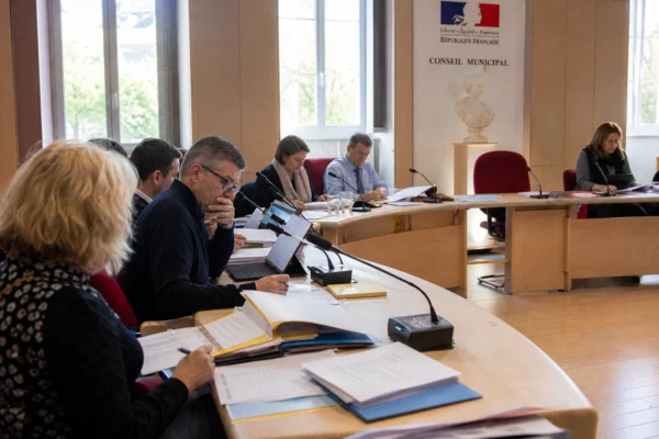 Salle de conseil municipal française avec une chaise vide, des micros et des dossiers budgétaires sur une table