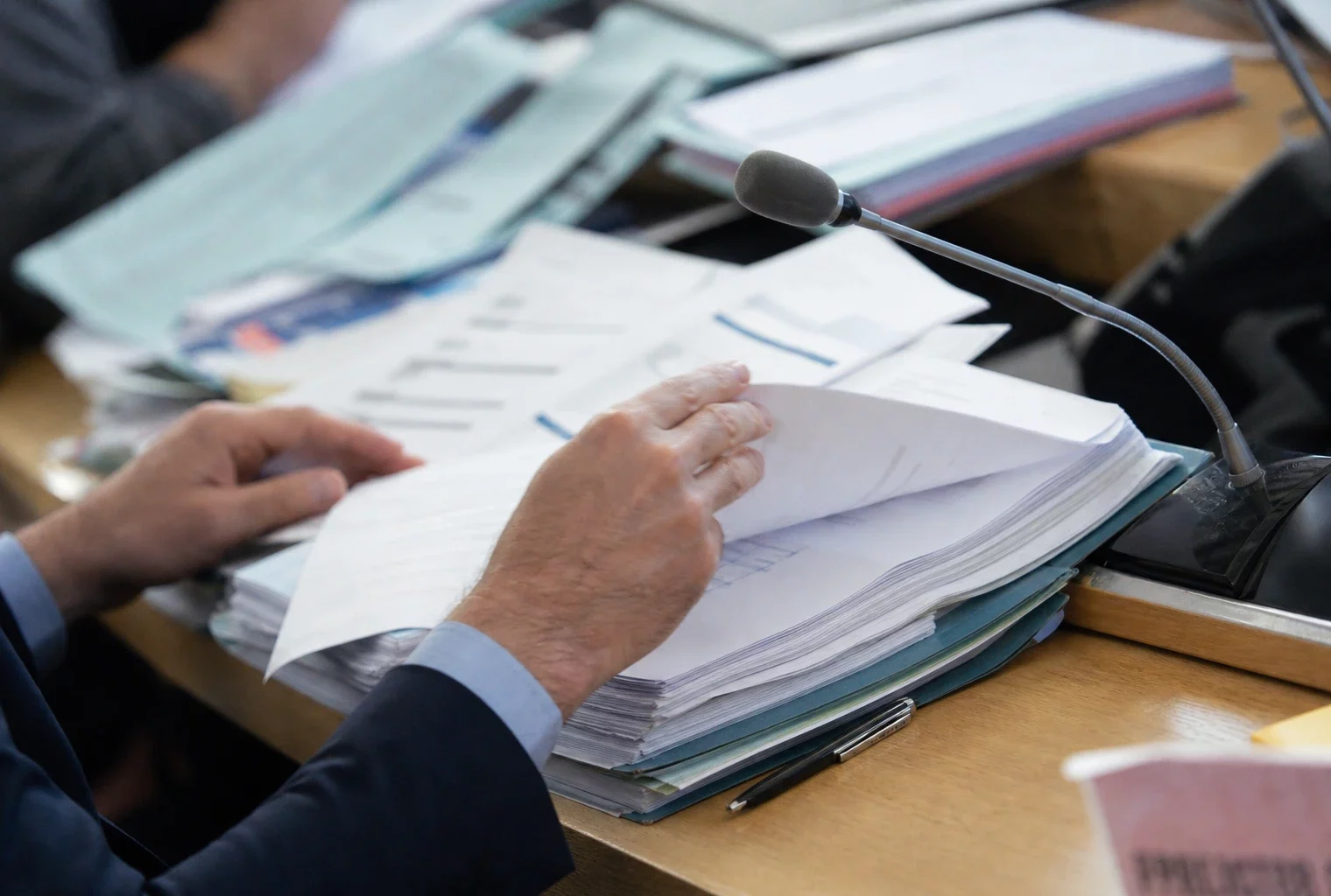 Dossier de commission budgétaire, mains anonymes, micro de commission et documents flous dans une lumière naturelle claire.
