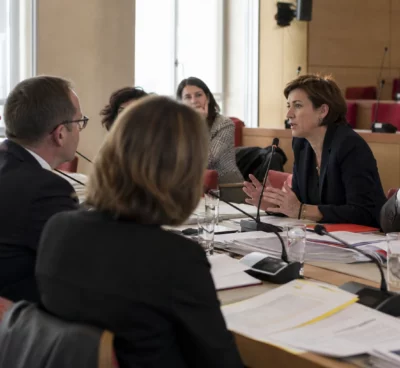 Réunion de commission parlementaire avec micros, dossiers ouverts et silhouettes anonymes dans une salle institutionnelle lumineuse
