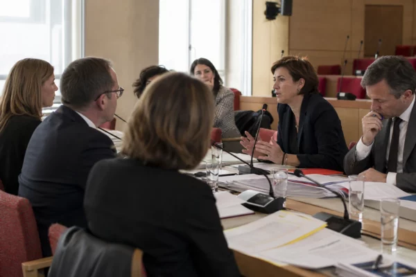 Réunion de commission parlementaire avec micros, dossiers ouverts et silhouettes anonymes dans une salle institutionnelle lumineuse