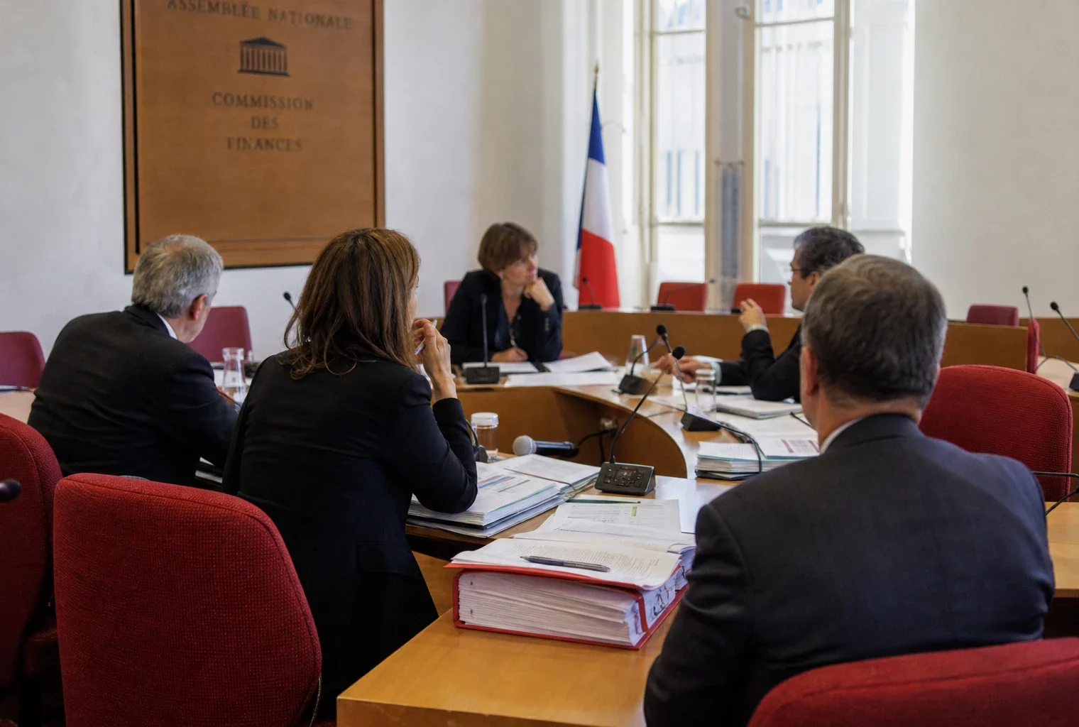 Réunion de commission parlementaire avec micros, dossiers ouverts et silhouettes anonymes dans une salle lumineuse