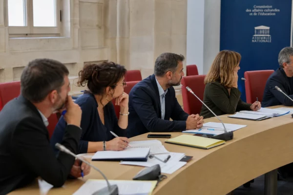Réunion de commission parlementaire sur l’école, avec enseignants anonymes, dossiers et microphones autour d’une table