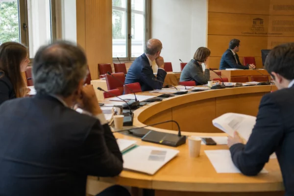 Réunion de commission parlementaire avec élus anonymes, micros et dossiers sous une lumière naturelle claire