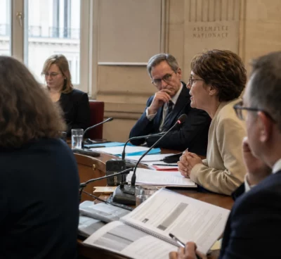 Réunion de commission parlementaire en France avec micros, dossiers ouverts et silhouettes anonymes autour d’une audition