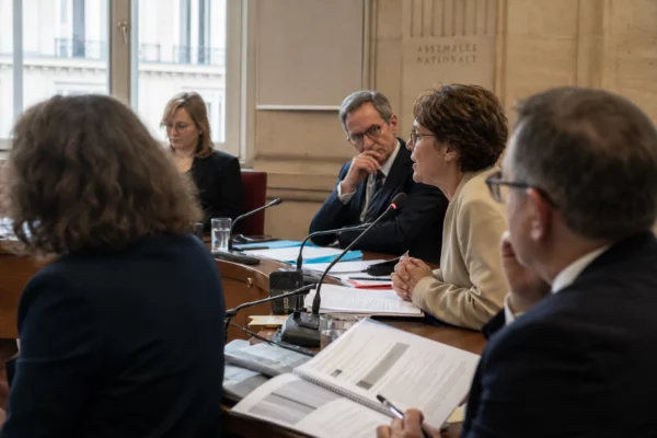 Réunion de commission parlementaire en France avec micros, dossiers ouverts et silhouettes anonymes autour d’une audition