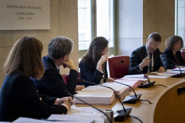 Réunion de commission parlementaire en France avec micros, dossiers ouverts et silhouettes anonymes sous une lumière claire.