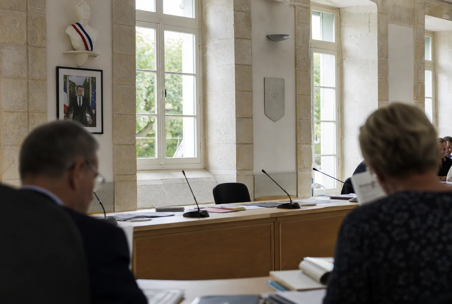 Salle municipale claire avec une chaise vide, des micros et des dossiers flous sur une table de conseil