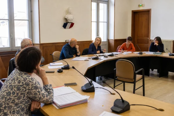 Conseil municipal français réuni autour d’une table, avec une chaise vide, des micros et des dossiers flous
