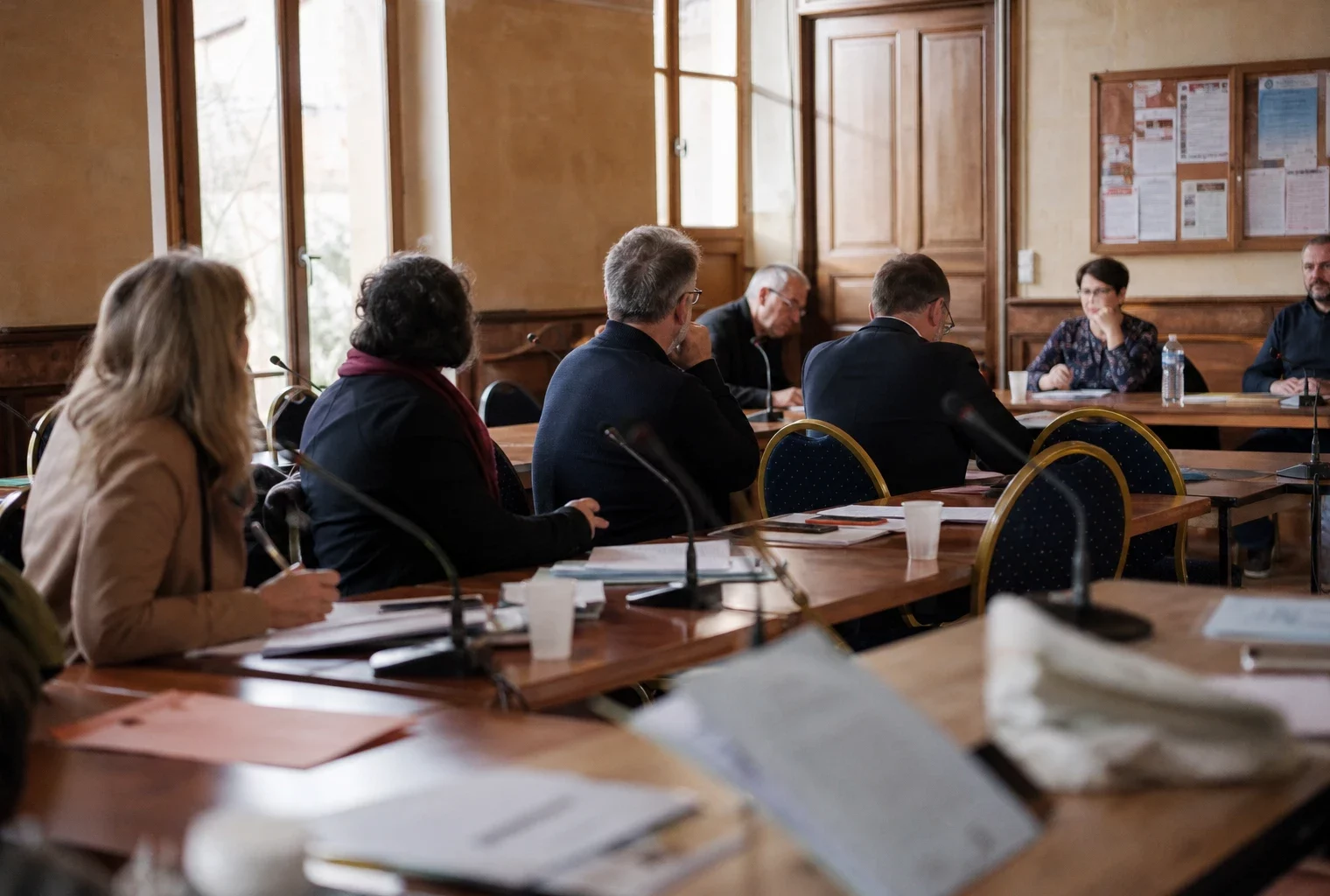 Salle de conseil municipal française avec une chaise vide, des micros et des dossiers autour d’une table