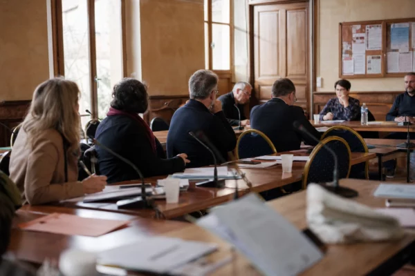 Salle de conseil municipal française avec une chaise vide, des micros et des dossiers autour d’une table