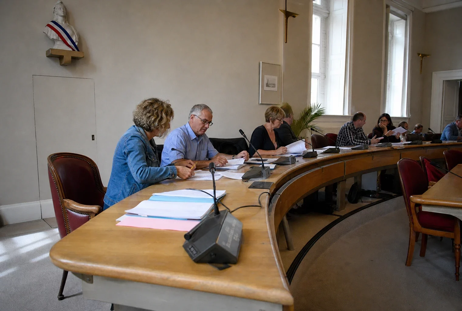 Salle municipale française avec élus anonymes, chaise vide, micros et dossiers lors d’un conseil local