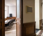 Couloir clair menant à une salle d’audition institutionnelle vide, avec porte entrouverte et micros sur table.