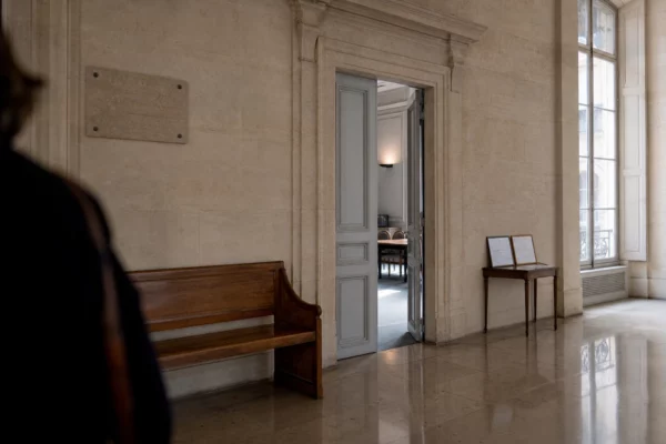 Couloir lumineux d’une institution française, porte entrouverte et salle d’audition visible au fond.