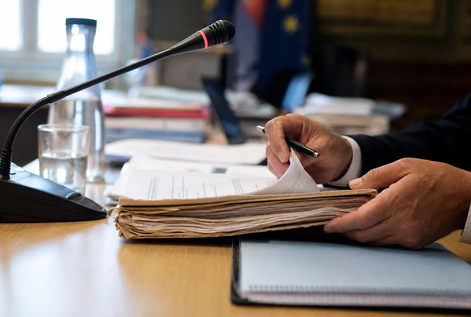 Mains anonymes feuilletant un dossier devant un micro de commission dans une salle institutionnelle lumineuse.