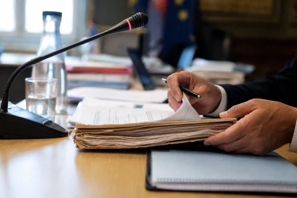 Mains anonymes feuilletant un dossier devant un micro de commission dans une salle institutionnelle lumineuse.