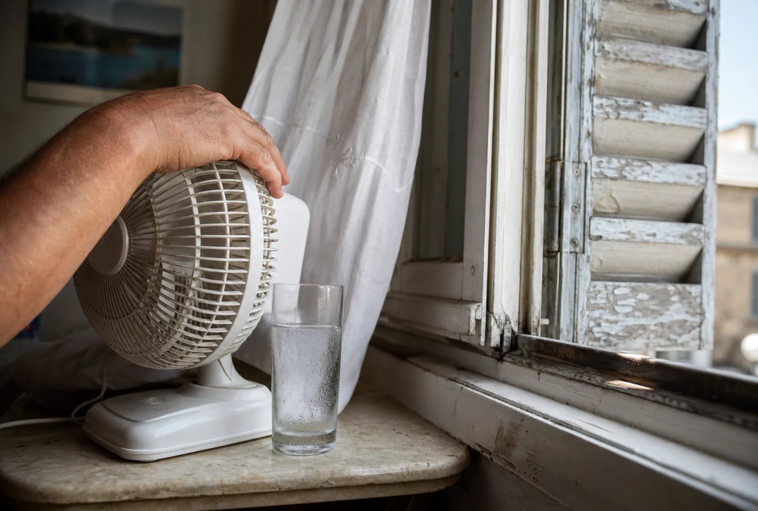 Main près d’un petit ventilateur dans un appartement français en canicule, avec un verre d’eau et des volets entrouverts