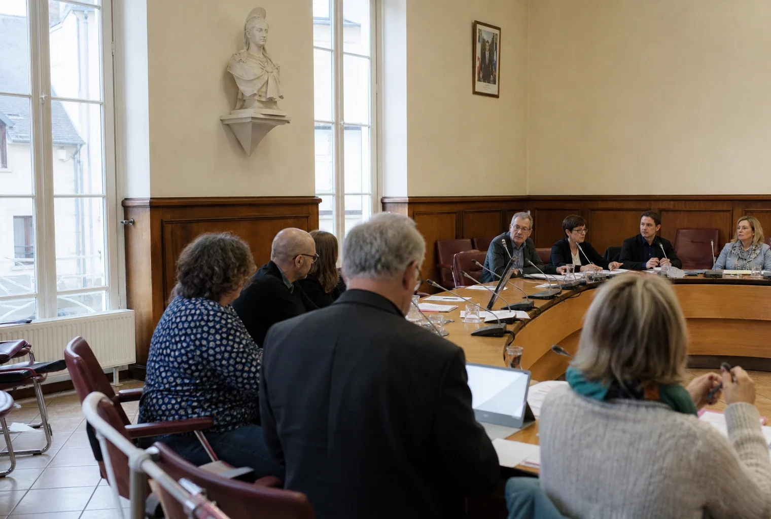 Salle de conseil municipal française avec une chaise vide, des micros et des dossiers autour d’une table