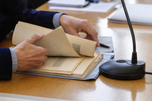 Mains anonymes examinant un dossier d’audition près d’un micro de commission, dans une lumière naturelle claire