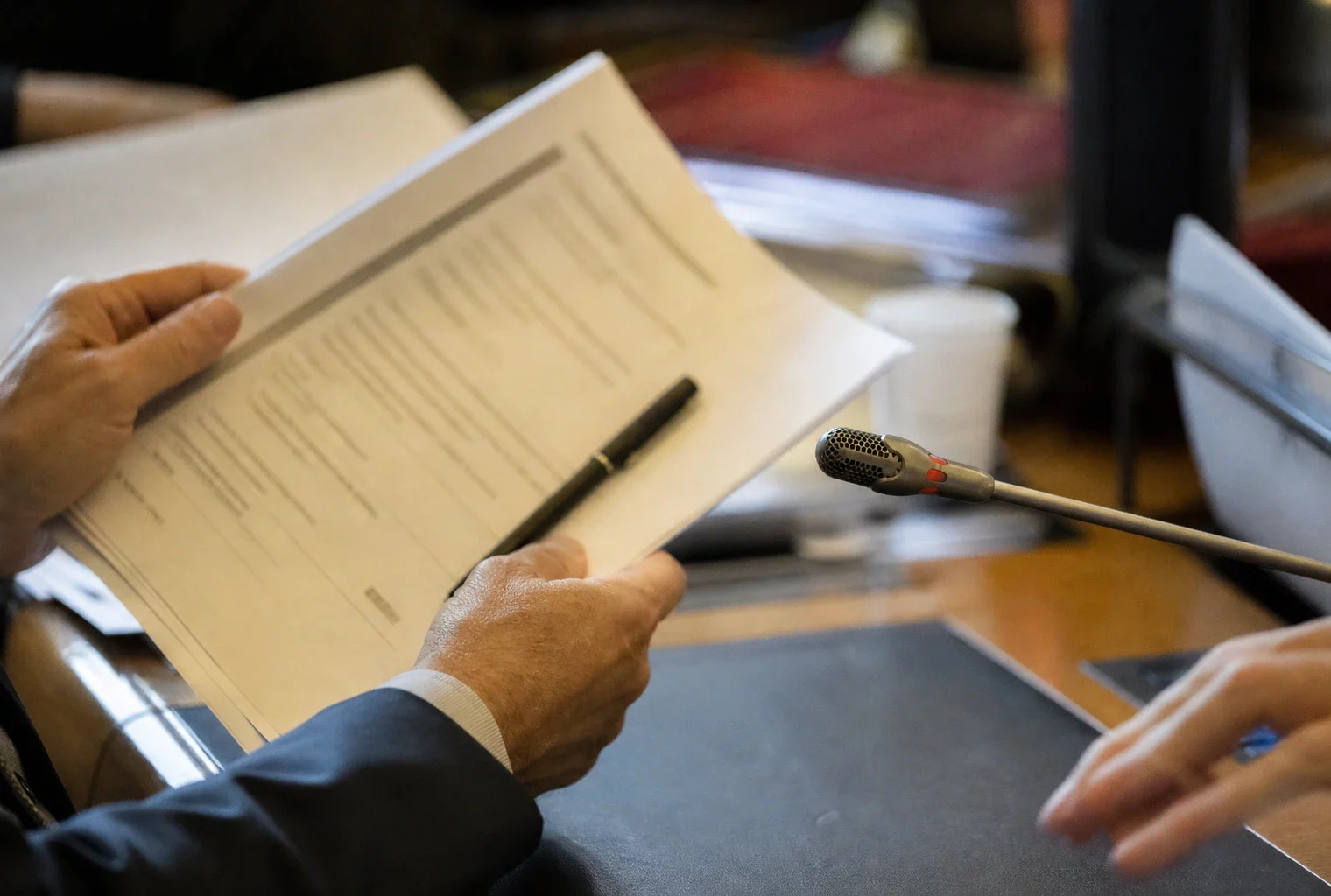 Mains anonymes tenant un dossier lors d’une audition parlementaire, avec un microphone et des documents flous