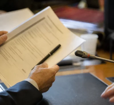 Mains anonymes tenant un dossier lors d’une audition parlementaire, avec un microphone et des documents flous