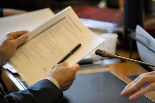 Mains anonymes tenant un dossier lors d’une audition parlementaire, avec un microphone et des documents flous