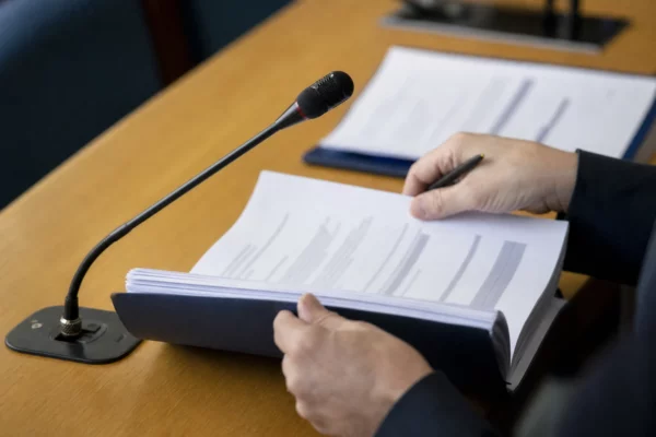 Mains anonymes examinant un dossier lors d’une audition parlementaire, avec un micro de commission et des documents flous