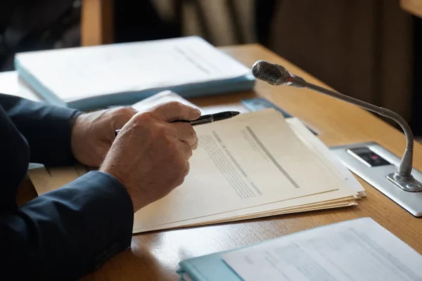 Mains anonymes examinant un dossier d’audition près d’un micro de commission, dans une lumière naturelle claire
