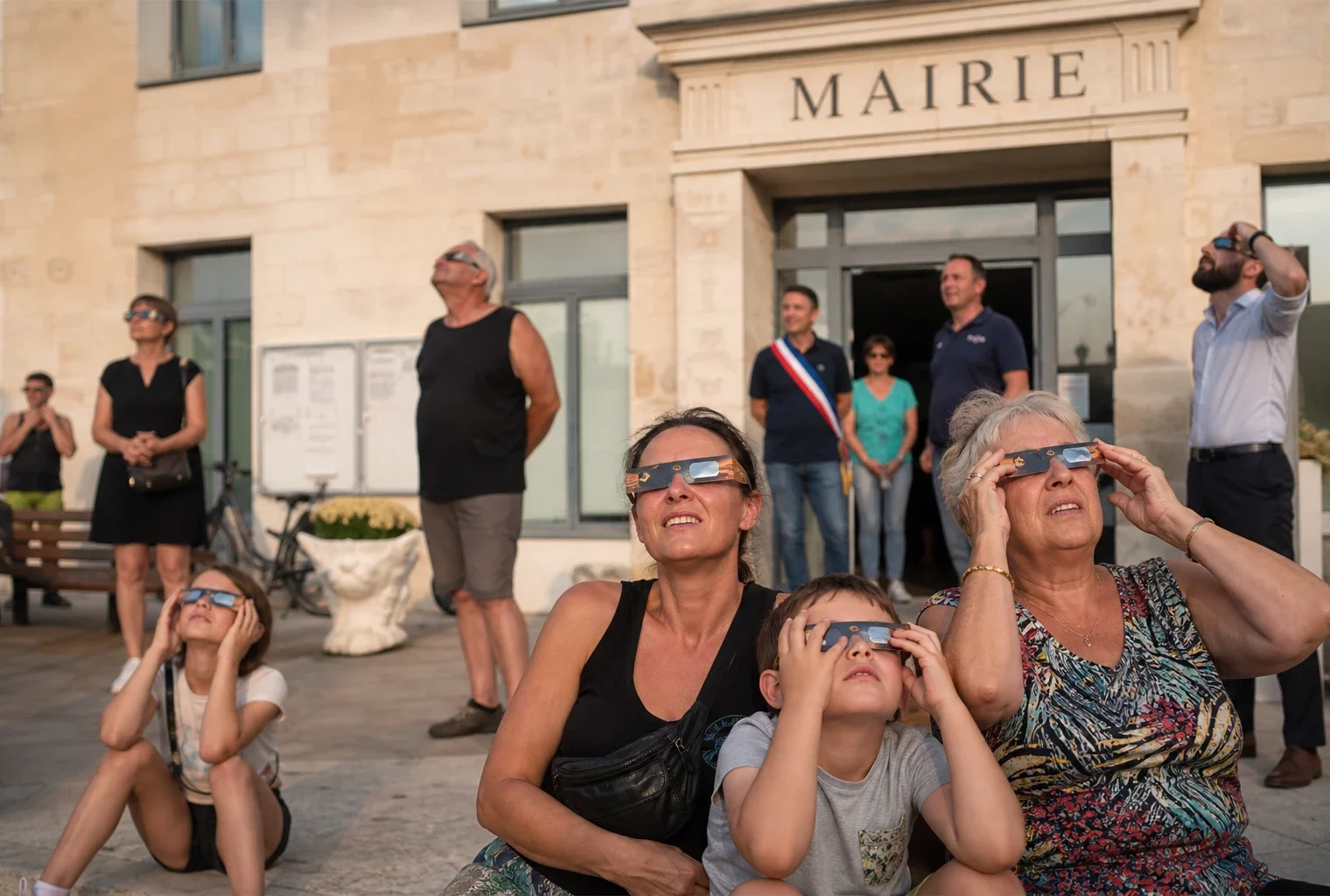 Des habitants observent une éclipse solaire partielle sur le parvis d’une mairie de village, au coucher du Soleil.
