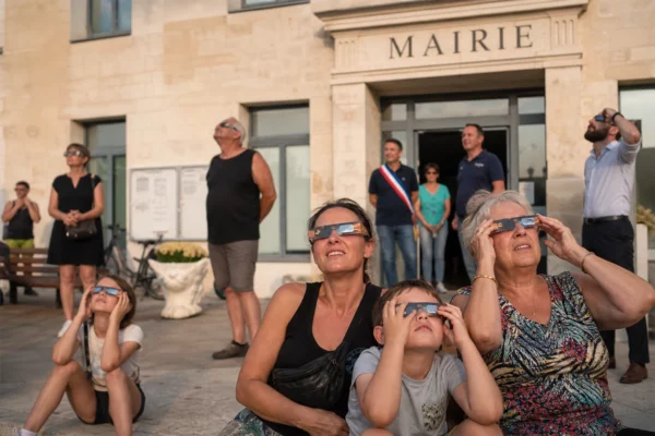 Des habitants observent une éclipse solaire partielle sur le parvis d’une mairie de village, au coucher du Soleil.