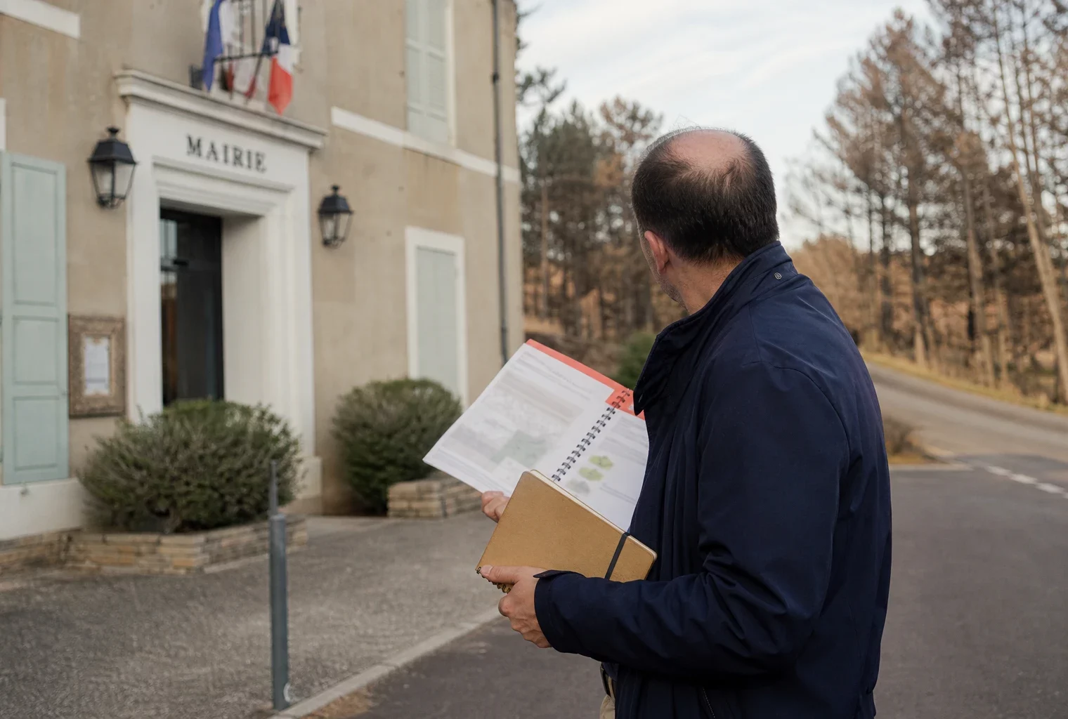 Élu local anonyme avec dossier de prévention incendie devant une mairie de village, en lumière naturelle.