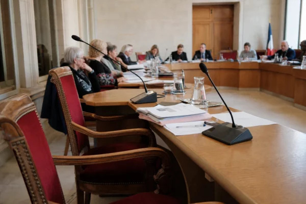 Salle municipale de conseil local avec micros, dossiers et chaise vide sur fond de réunion calme.