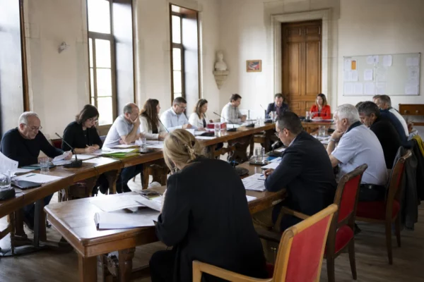 Salle de conseil municipal française avec micros, dossiers flous et une chaise vide pendant une réunion politique